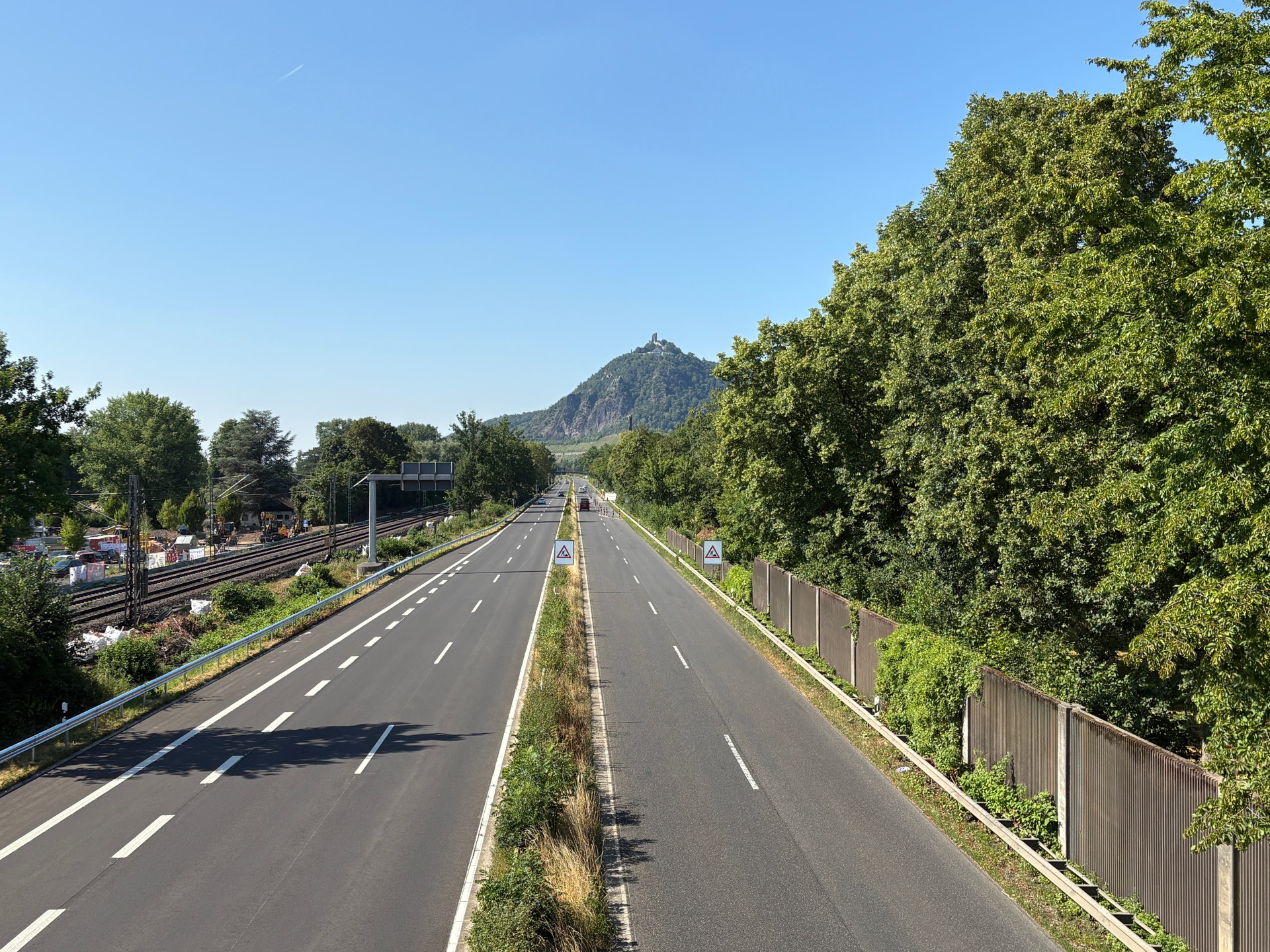 **Alt-Text:** Blick auf die B 42 bei Bad Honnef mit den Bahngleisen und einer Baustelle links sowie dem Drachenfels im Hintergrund.