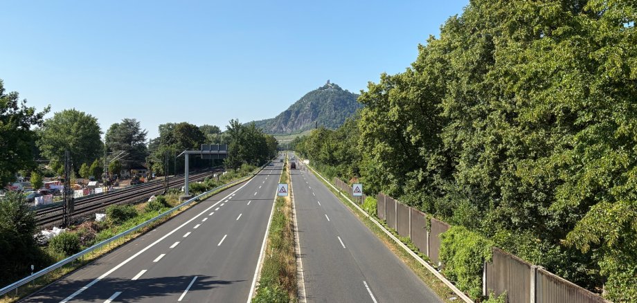 **Alt-Text:** Blick auf die B 42 bei Bad Honnef mit den Bahngleisen und einer Baustelle links sowie dem Drachenfels im Hintergrund.
