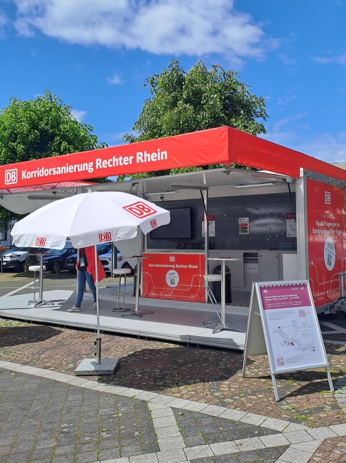 Infomobil der Deutschen Bahn zur Korridorsanierung der rechtsrheinischen Bahnstrecke. Der rote Informationsstand mit der Aufschrift „Korridorsanierung Rechter Rhein“ steht auf einem Platz im Freien. Vor dem Pavillon befinden sich Stehtische, Sonnenschirme mit DB-Logo sowie eine Informationstafel für Besucherinnen und Besucher.