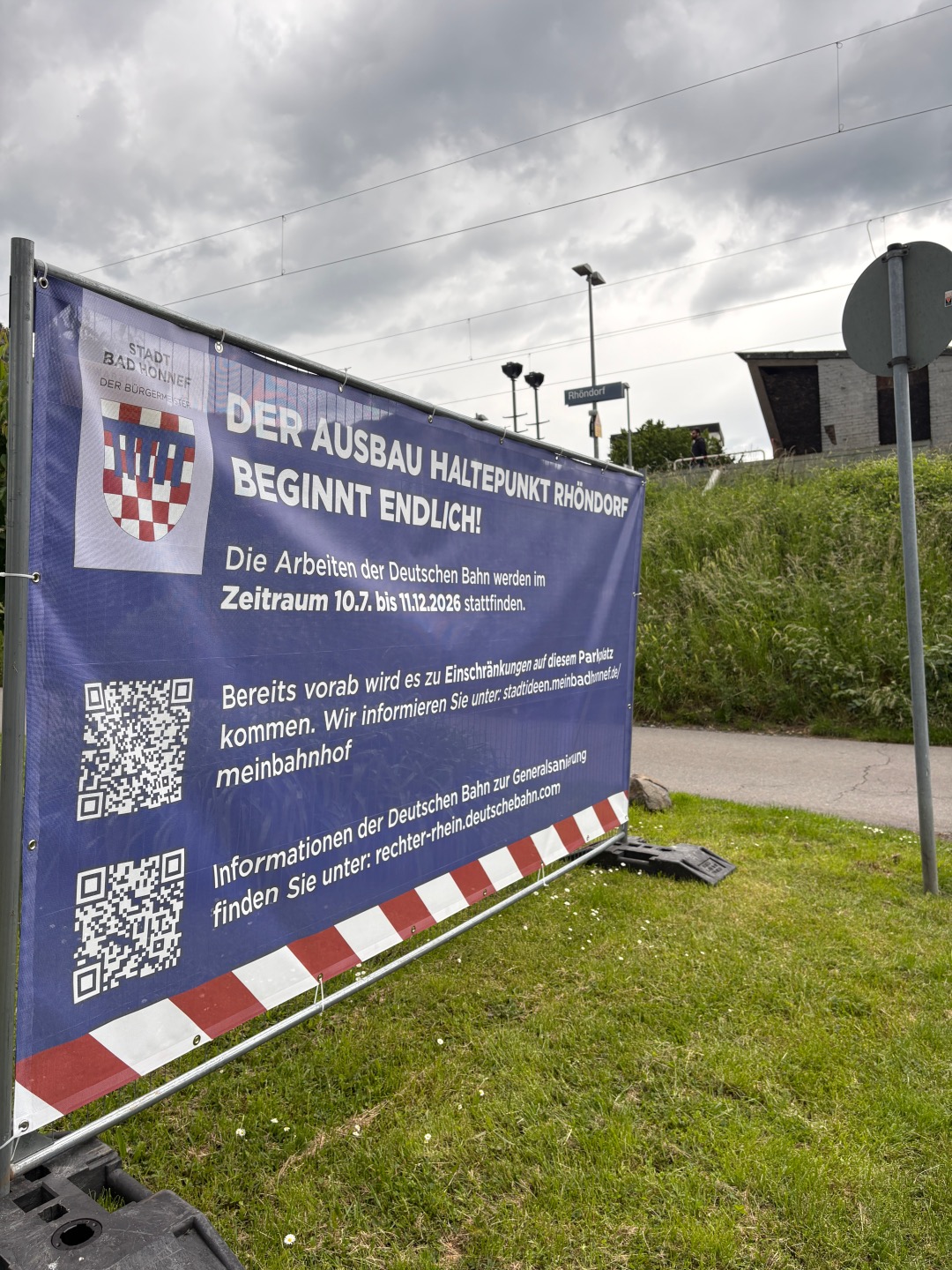 Großes Informationsbanner der Stadt Bad Honnef am Bahnhaltepunkt Rhöndorf mit Hinweis auf den Ausbau des Haltepunktes und bevorstehende Einschränkungen während der Bauarbeiten. Im Hintergrund sind Bahnhofsbereich und bewölkter Himmell zu sehen.