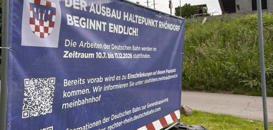 Großes Informationsbanner der Stadt Bad Honnef am Bahnhaltepunkt Rhöndorf mit Hinweis auf den Ausbau des Haltepunktes und bevorstehende Einschränkungen während der Bauarbeiten. Im Hintergrund sind Bahnhofsbereich und bewölkter Himmell zu sehen.