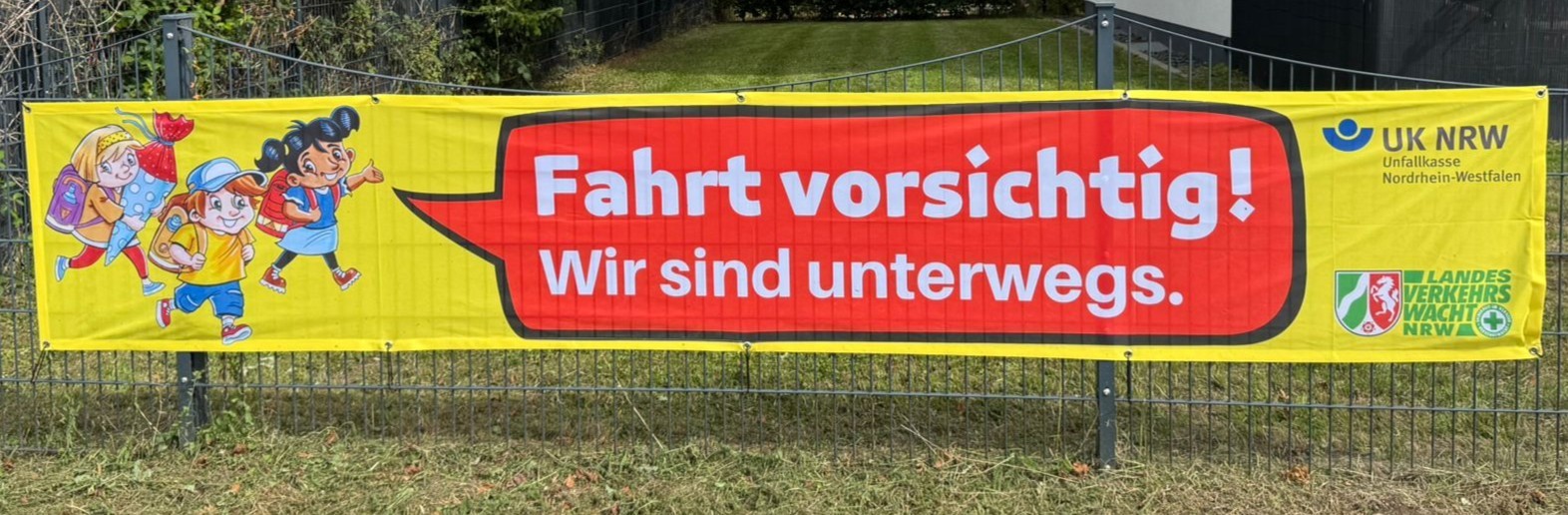 Auf einem Banner ist eine Gruppe Schulkinder zu sehen, die die Verkehrsteilnehmenden  bitten "Fahrt vorsichtig. Wir sind unterwegs!"