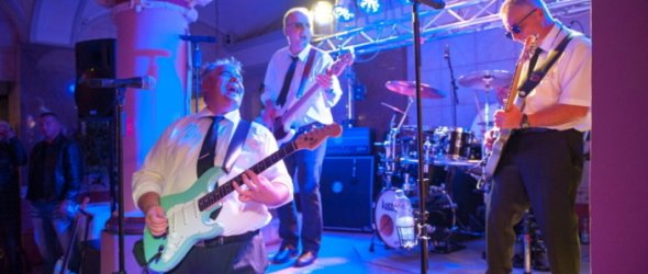 Live-Band spielt mit Gitarren und Bass auf einer beleuchteten Bühne bei einer Abendveranstaltung, Publikum im Hintergrund
