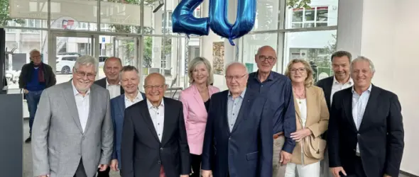 Gruppenfoto von festlich gekleideten Menschen in einem Foyer, blaue Ballons mit der Zahl 20 hängen über ihnen.