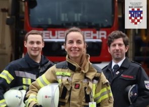 Drei Feuerwehrkräfte stehen vor einem Einsatzfahrzeug und lächeln in die Kamera, zwei mit Einsatzkleidung und Helmen in der Hand.