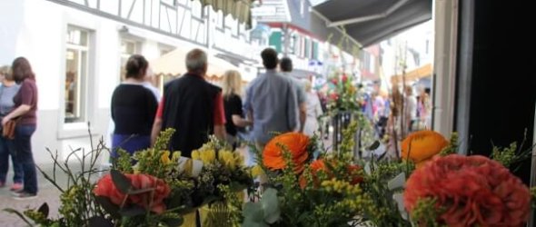 Bunte Blumensträuße im Vordergrund, dahinter Menschen, die durch eine belebte Altstadtstraße schlendern.