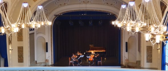 Musiker auf einer Bühne in einem historischen Festsaal mit Kronleuchtern, Publikum sitzt im unteren Bereich.