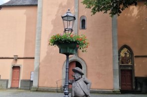 Steinerne Brunnenfigur neben einer Laterne mit Blumen, angestrahlte Kirchenfassade im Hintergrund.