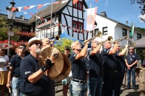 Blas- und Schlagmusikgruppe spielt im Freien unter bunten Wimpeln, Zuschauer stehen am Straßenrand.