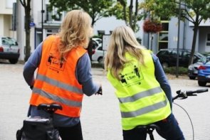 Zwei Radfahrende tragen gut sichtbare Warnwesten und stehen mit Fahrrädern auf einem Platz in der Stadt.