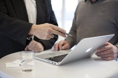 Zwei Personen stehen an einem runden Tisch und besprechen etwas an einem aufgeklappten Laptop, ein Glas Wasser und Visitenkarten liegen daneben