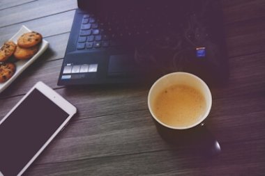 Arbeitsplatz mit Laptop, Tasse Kaffee, Smartphone und einem Teller mit Keksen auf einem Holztisch.