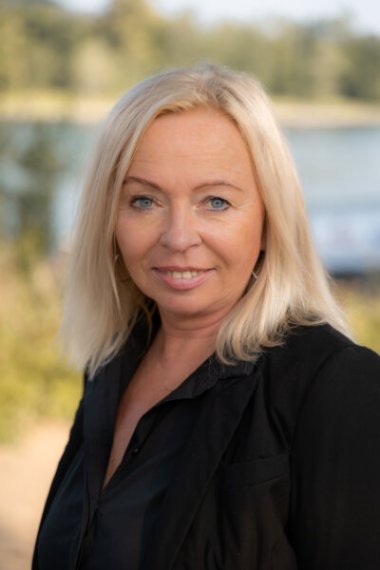 Andrea Enzenberger: Porträt einer Frau mit blondem Haar in dunkler Kleidung, sie lächelt in die Kamera, im Hintergrund ist eine Flusslandschaft erkennbar.