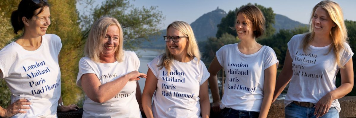 Team Wirtschaftsförderung: Fünf Frauen in weißen T-Shirts mit Aufdruck stehen lachend im Freien vor einer Fluss- und Bergkulisse und unterhalten sich.