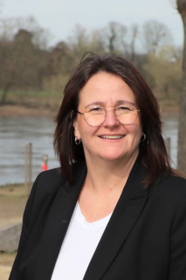Cornelia Jungheim: Porträt einer Frau mit dunklem Haar und Brille, sie lächelt in die Kamera, ein Fluss und Bäume sind im Hintergrund zu sehen.