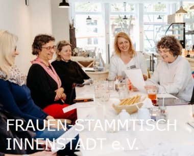 Gruppe von Frauen sitzt gemeinsam an einem Tisch in einem Café, sie unterhalten sich und lachen miteinander, Gläser und Schreibunterlagen liegen auf dem Tisch.