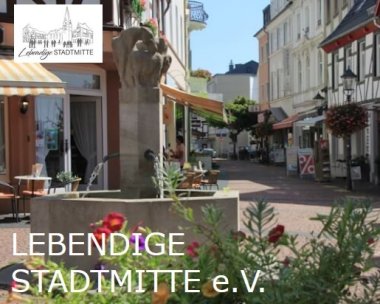 Gemütliche Fußgängerzone mit einem Brunnen im Vordergrund, Cafés und Blumenbeete rundherum, Sonnenschirme spenden Schatten.