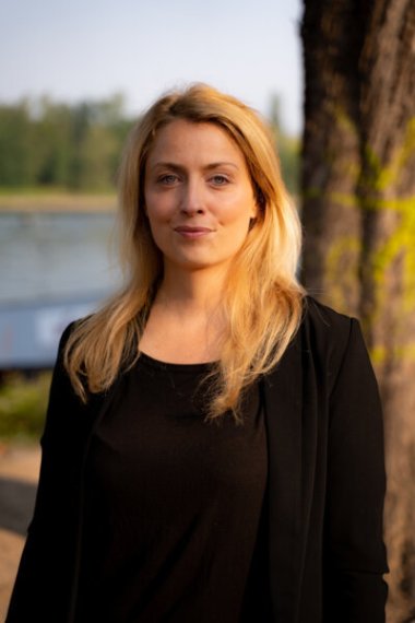 Miriam Brackelsberg Miriam Brackelsberg: Porträt einer Frau mit langen blonden Haaren, sie steht vor einem Baum und einem Flussufer und blickt ruhig in die Kamera.