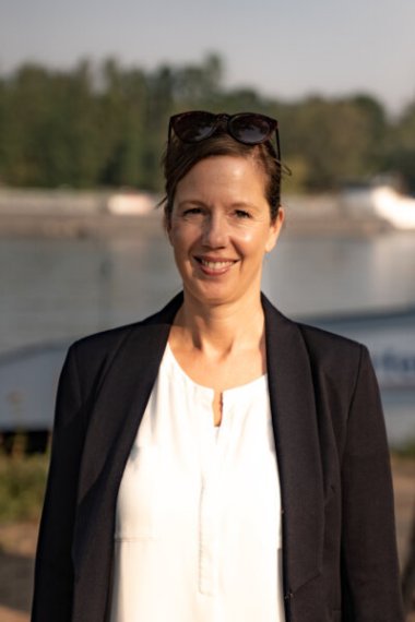 Sonja Schwalbe Sonja Schwalbe: Porträt einer Frau mit zusammengebundenem Haar und dunklem Blazer, sie lächelt und steht vor einem Flussufer