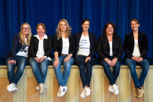 Team Wirtschaftsförderung: Sechs Frauen sitzen nebeneinander vor einem blauen Bühnenvorhang, alle tragen Jeans und dunkle Blazer und schauen lächelnd in die Kamera.