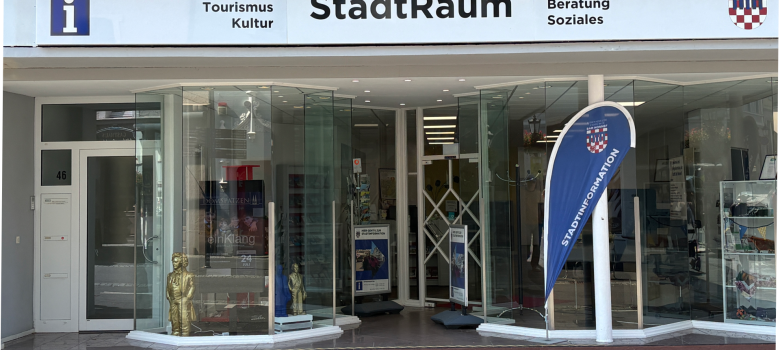 Außenansicht des StadtRaums Bad Honnef in der Hauptstraße 46 mit großer Glasfassade. Über dem Eingang stehen die Hinweise „Stadtinformation“, „Tourismus“, „Kultur“, „Ehrenamt“, „Beratung“ und „Soziales“. Vor dem Eingang befindet sich eine blaue Fahne der Stadtinformation.