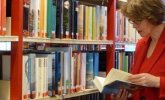 Regal mit vielen bunten Büchern. Daneben eine ältere Frau mit einem aufgeschlagenen Buch in der Hand