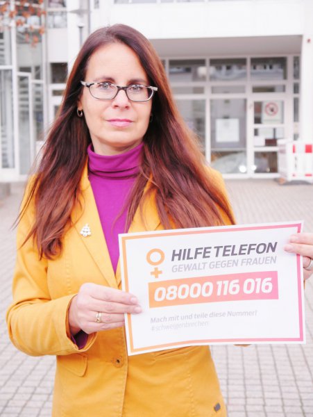 Iris Schwarz zeigt vor dem Rathaus das Schild mit der Nummer des Hilfetelefons
