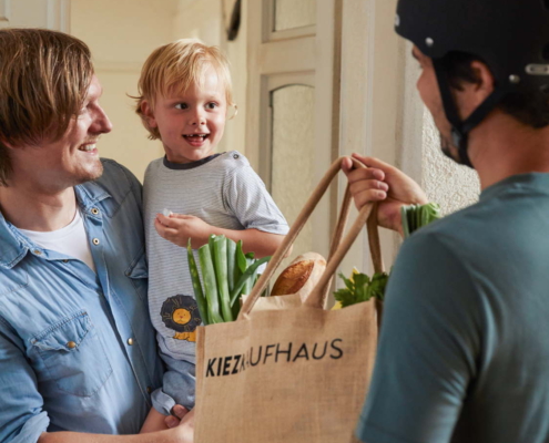 Ein Fahrradkurier überreicht einem Mann eine mit grünem Gemüse gefüllte Einkaufstasche aus Jute. Der Mann steht im Türrahmen seiner Wohnung und trägt sein Kind auf dem Arm. Das kleine Kind lächelt den Fahrradkurier an.