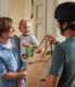 Ein Fahrradkurier überreicht einem Mann eine mit grünem Gemüse gefüllte Einkaufstasche aus Jute. Der Mann steht im Türrahmen seiner Wohnung und trägt sein Kind auf dem Arm. Das kleine Kind lächelt den Fahrradkurier an.