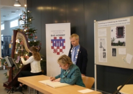 Sylvia Löhrmann sitzend beim Eintrag ins Goldene Buch, Bürgermeister Otto Neuhoff stehend, Harfenspielerin