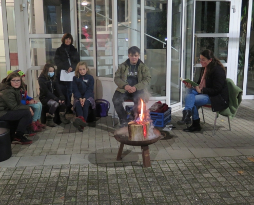 Kinder sitzen auf Bänken vor dem Rathaus Bad Honnef einer Feuerschale. Alle tragen warme Winterkleidung. Es ist fast dunkel, nur die kleine Flamme leuchtet hell. Eine erwachsene Frau liest ihnen eine Episode aus dem Kinderbuch "Ronja Räubertochter" von Astrid Lindgren vor.