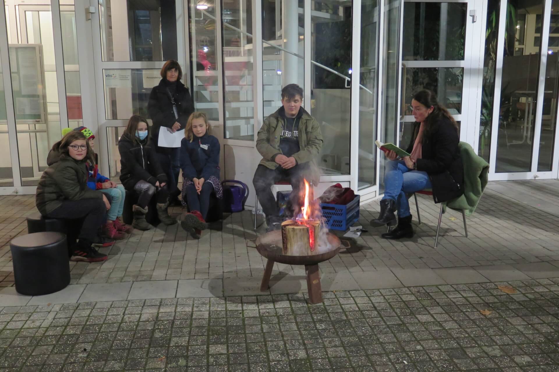 Kinder sitzen auf Bänken vor dem Rathaus Bad Honnef einer Feuerschale. Alle tragen warme Winterkleidung. Es ist fast dunkel, nur die kleine Flamme leuchtet hell. Eine erwachsene Frau liest ihnen eine Episode aus dem Kinderbuch "Ronja Räubertochter" von Astrid Lindgren vor.