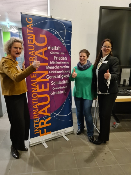 Frauke Fischer, Johanna Samaras und Iris Schwarz vor dem Roll-up zum Internationalen Frauentag mit hochgestellten Daumen 